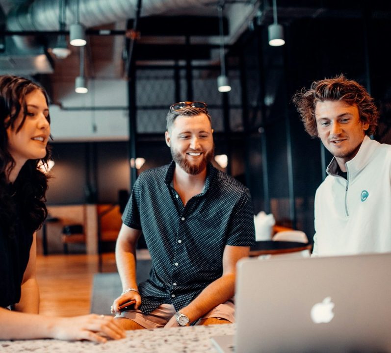 Three people engaged in a discussion around a laptop in a modern workspace.