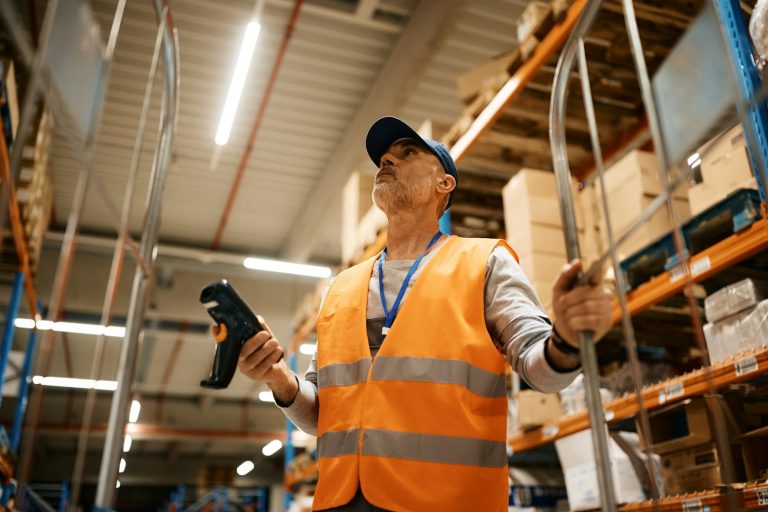 Warehouse worker in high-visibility vest holding a scanner, surrounded by shelves of goods.