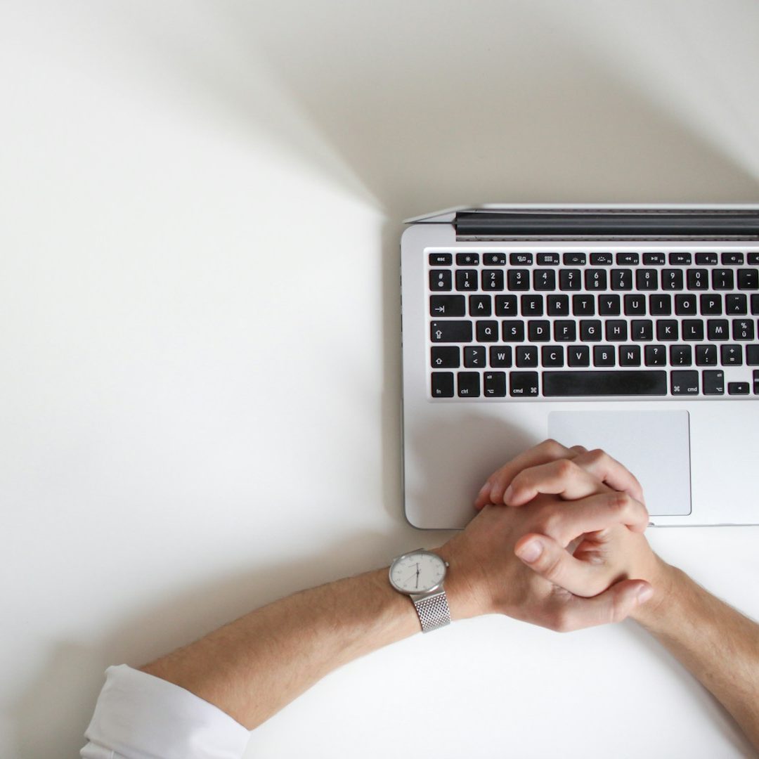 A pair of hands resting on a desk beside a laptop.