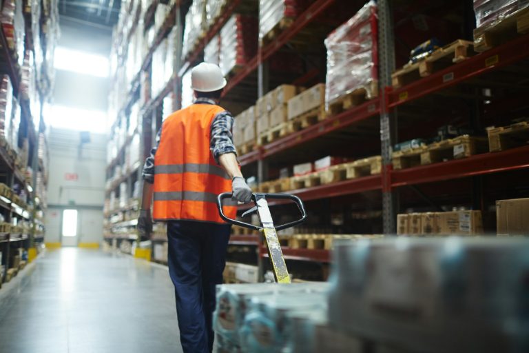 Warehouse worker in high-visibility vest using a pallet jack among shelves of goods.